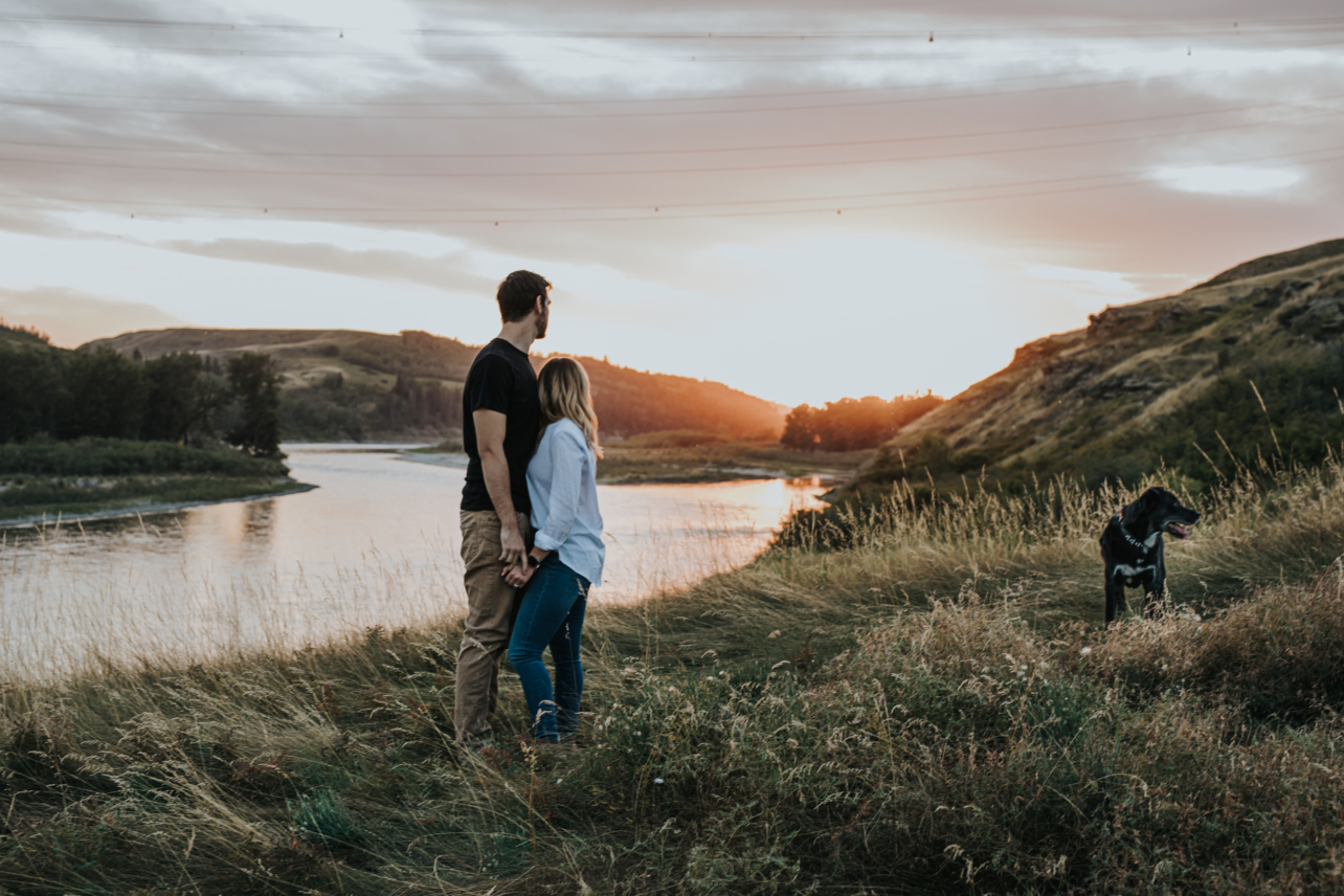 Couple embraced watching sunset during summer engagement session with black dog nearby